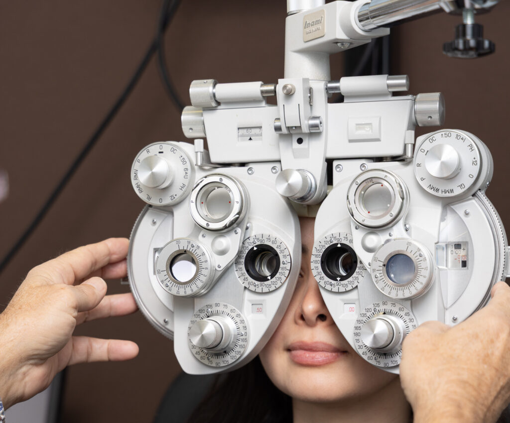 A woman is sitting for an eye test while an optometrist adjusts a phoropter in front of her face to test her vision.