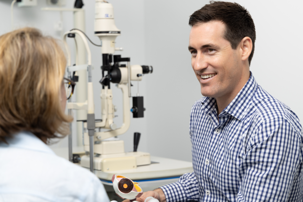 A smiling optometrist in a chequered shirt holds an eye model whilst talking to a patient in a Gold Coast examination room on Nerang Street, with eye testing equipment in the background.