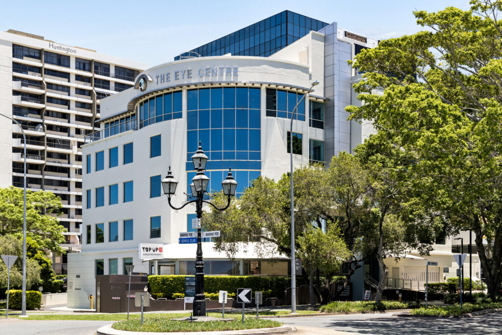 A modern, multi-storey building labelled The Eye Centre with glass windows, surrounded by trees and street signs, stands on a sunny day—home to leading Laser Eye Surgery Gold Coast services, with other office buildings in the background.