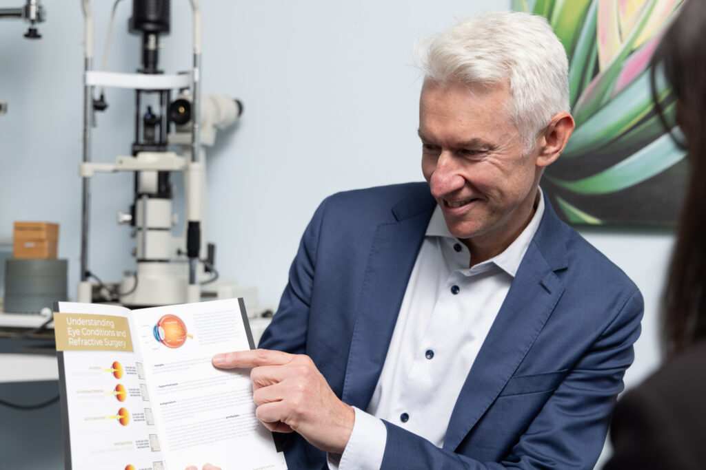 A man in a blue suit is smiling and pointing at an informational eye chart in a medical surgery, explaining eye conditions to a person sitting opposite him. Eye examination equipment is visible in the background.