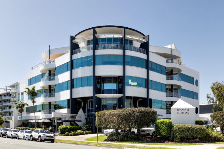 A modern, four-storey office building with blue-tinted windows and rounded architectural features on a sunny day. Several cars are parked along the street near a white sign that reads Esplanade & Second, home to Lasersight Sunshine Coast.