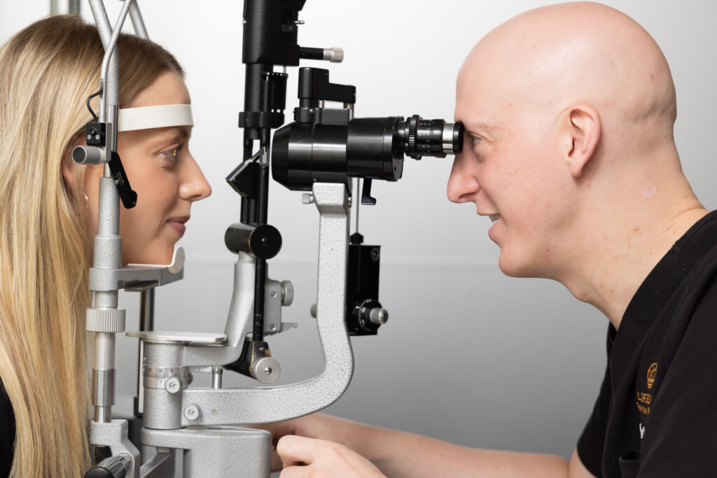 A woman sits at an eye examination machine while an eye care professional looks through the device to examine her eyes in a clinical setting on the Sunshine Coast.