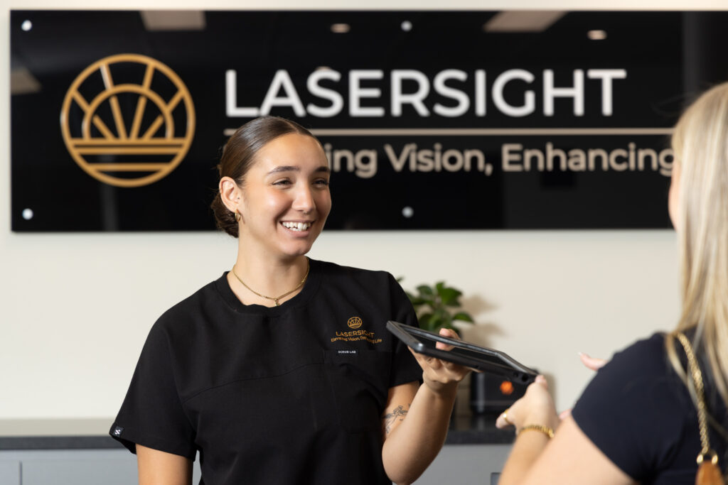 A smiling receptionist in a black uniform hands a tablet to a visitor at the front desk of LASERSIGHT’s Sunshine Coast clinic, with the clinic’s logo and slogan visible on a sign in the background.