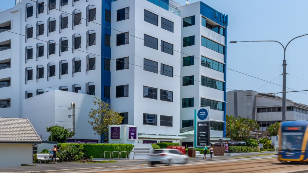 A modern white multi-storey medical centre with blue accents stands by Nerang Street on the Gold Coast. A tram passes in the foreground, with signs, greenery, and a few people near the building entrance.