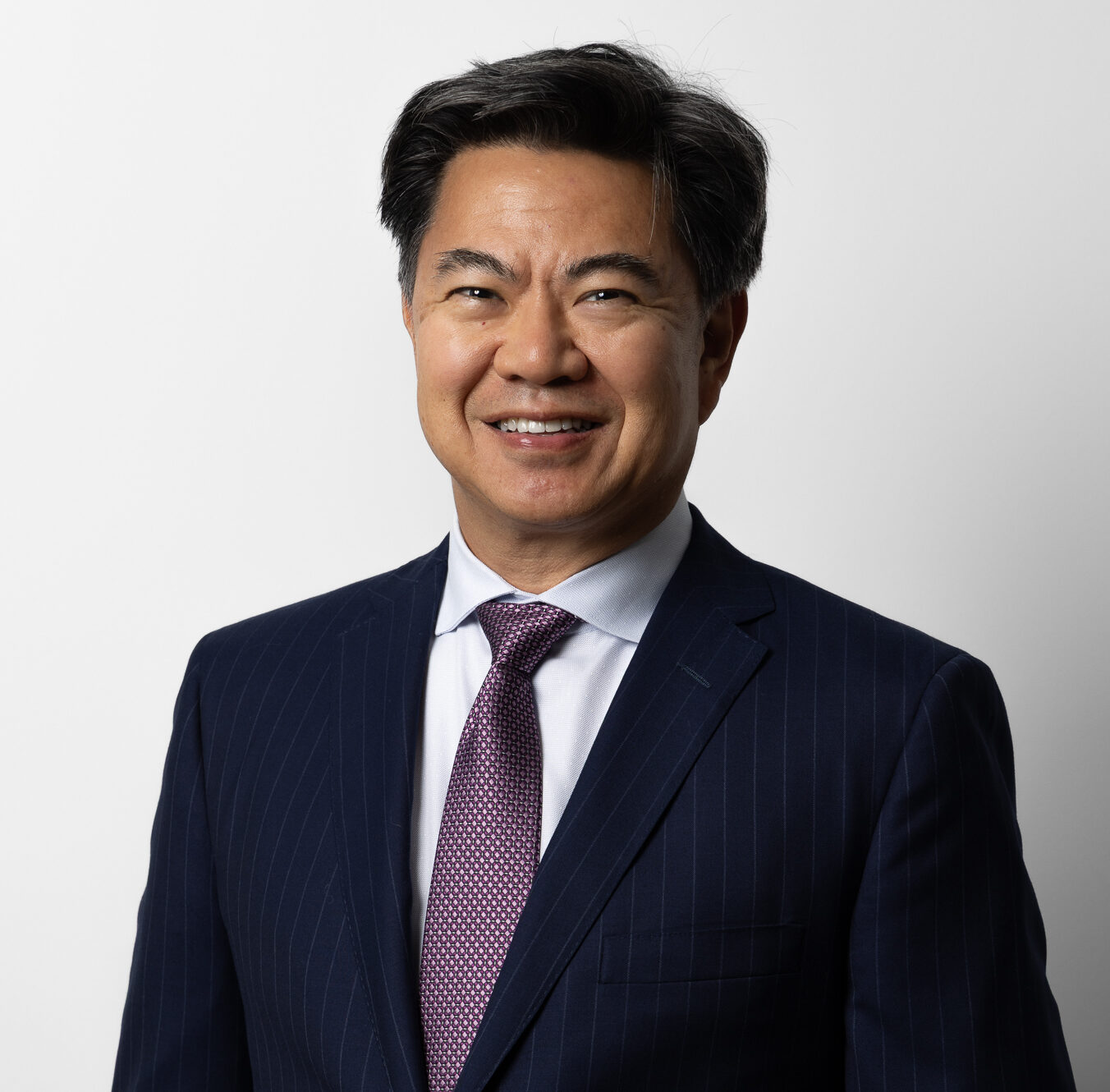 Dr Marc Wei in a navy blue pinstripe suit, white shirt, and purple tie stands smiling in front of a plain white background.