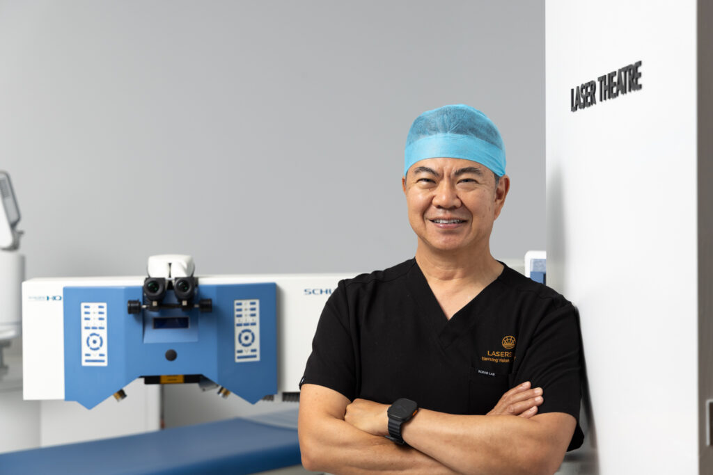 A smiling surgeon in black scrubs and a blue hair cap stands with arms crossed in a modern clinic, next to a sign reading LASER THEATRE and a large medical laser machine.