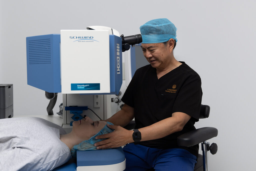 Dr Marc Wei wearing surgical scrubs and a blue cap positions a patient under a laser eye surgery machine in a medical clinic. The patient lies still with a protective cap on their head.