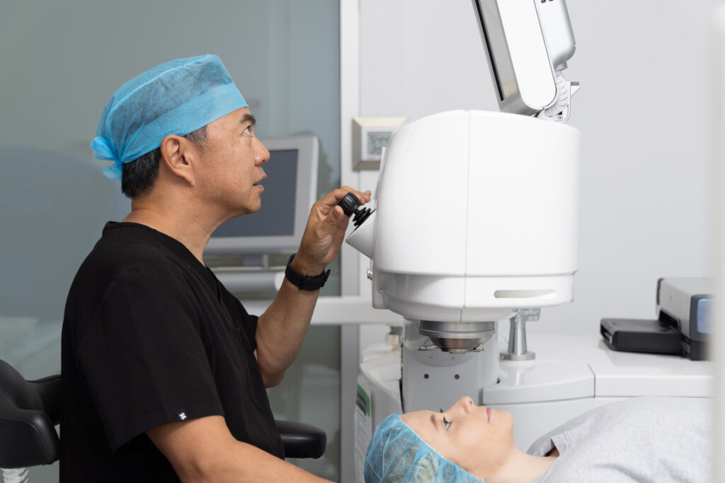 Dr Marc Wei in scrubs and a blue hair cover operates a laser eye surgery machine whilst a patient, also in a hair cover, lies on the procedure bed beneath the device.