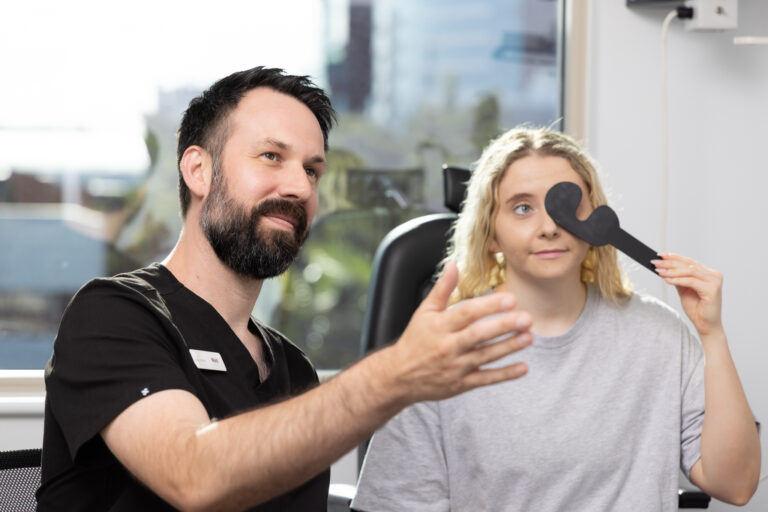 A bearded optometrist gestures while a blonde woman covers one eye with an eye occluder during an eye test in a bright clinic room on Short Street, Gold Coast.