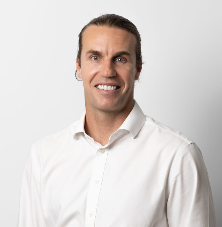 Dr Brett Drury, a man with long brown hair tied back and wearing a white button-up shirt, smiles at the camera against a plain white background.