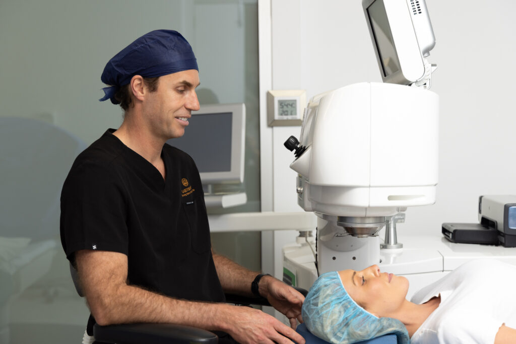 Dr Brett Drury, a medical professional wearing scrubs and a blue cap, prepares a patient for an eye procedure using a large ophthalmic machine in a clinical setting.