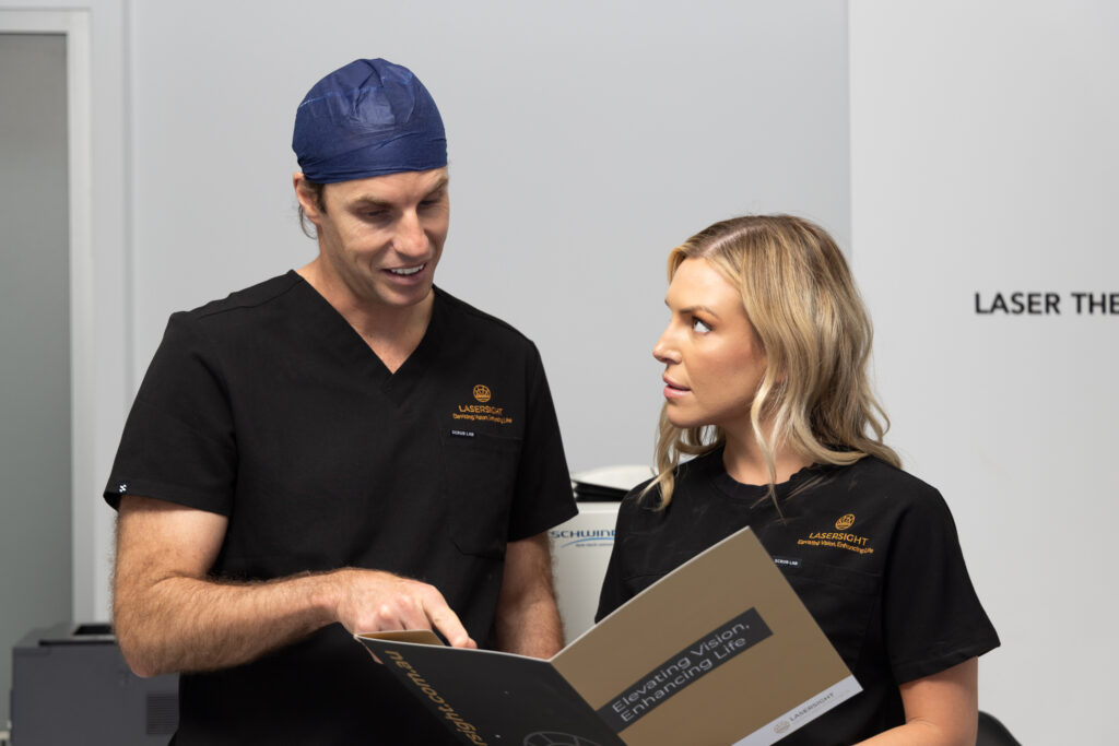 Two medical professionals in black scrubs, including Dr Brett Drury, discuss information from a booklet in a clinical setting. One holds the booklet open whilst the other listens attentively. Both appear focused and engaged in conversation.
