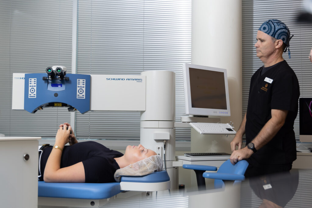 A patient lies on a medical bed under an eye surgery laser machine as Dr Hamish McKee, dressed in theatre scrubs and a surgical cap, stands nearby monitoring the procedure in a modern clinic.