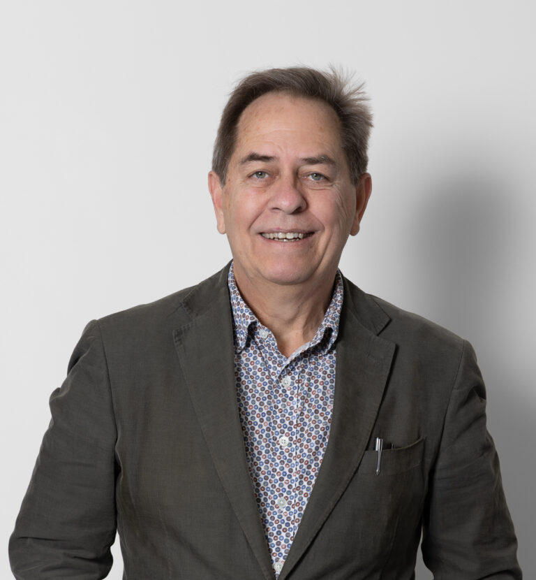 Dr Laurence Sullivan, a middle-aged man with short brown hair, is smiling at the camera. He is wearing a dark blazer over a patterned shirt, hands in his pockets, standing against a plain white background.
