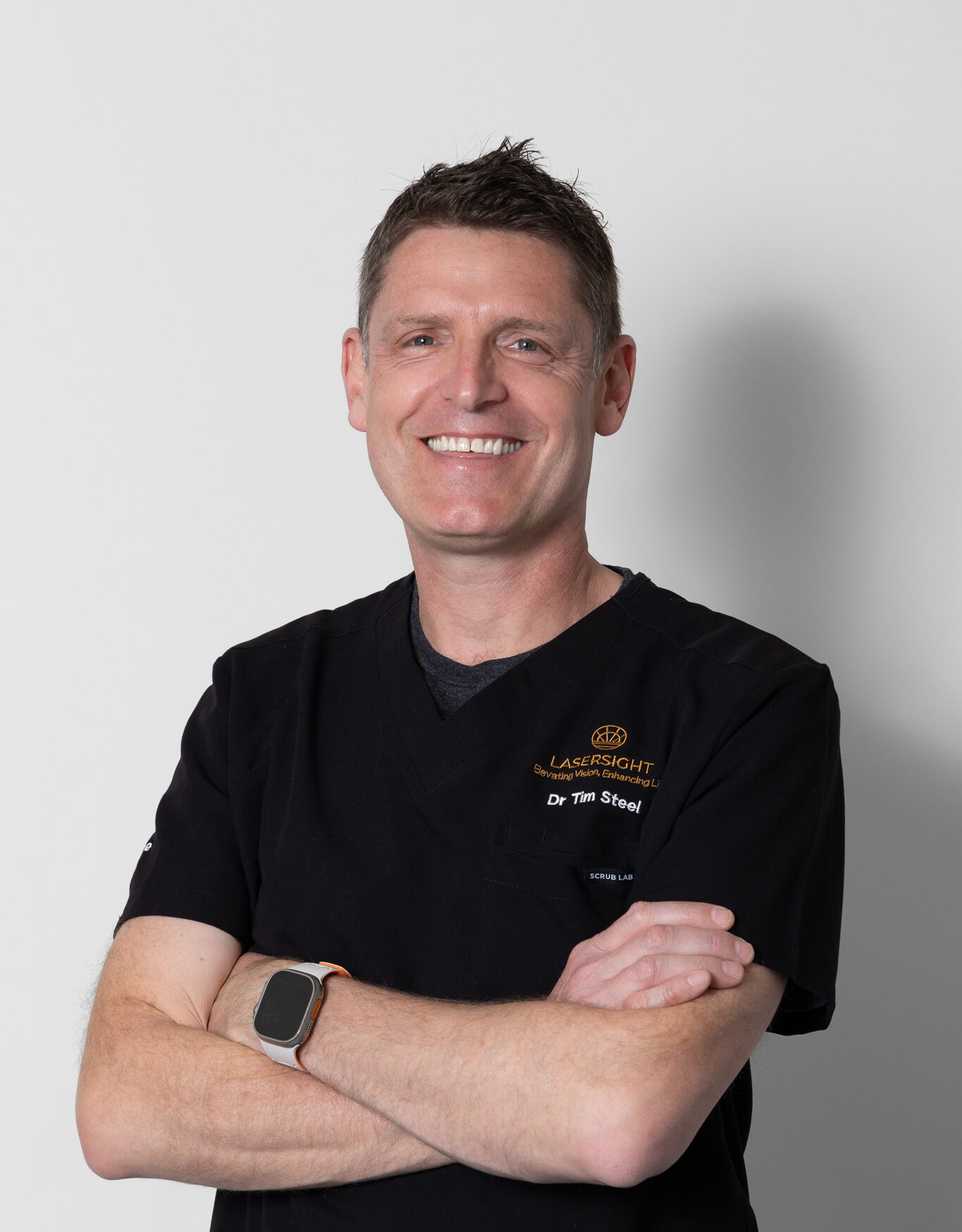 Dr Tim Steele, a smiling man with short brown hair, stands against a plain white background, arms folded. He wears black medical scrubs with embroidered text and a smart watch on his left wrist.