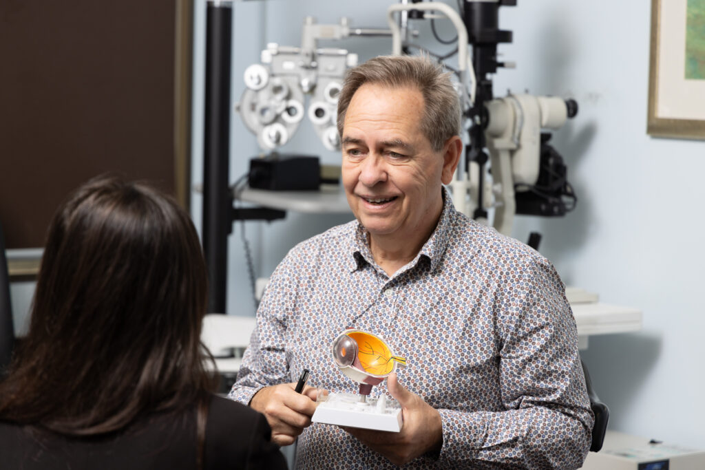 Dr Laurence Sullivan, a male optometrist, holds an anatomical eye model as he explains vision to a patient in an eye examination room, with eye examination equipment visible in the background.