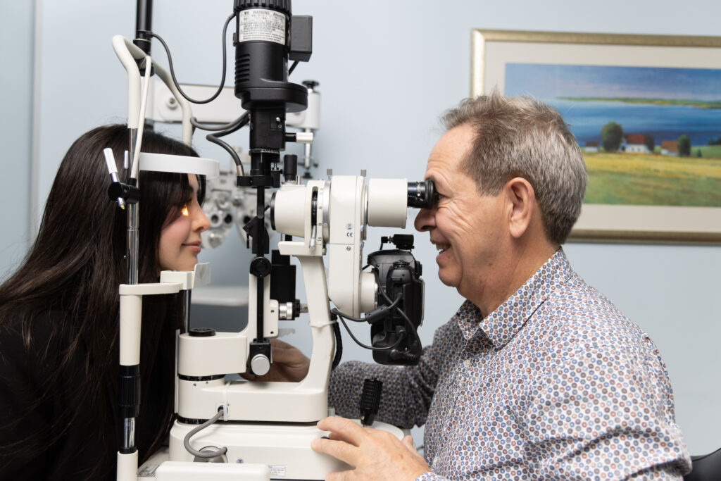 Dr Laurence Sullivan, a female optometrist, examines an older man’s eyes using a slit lamp in an eye clinic. The man is smiling, and a landscape painting hangs on the wall in the background.