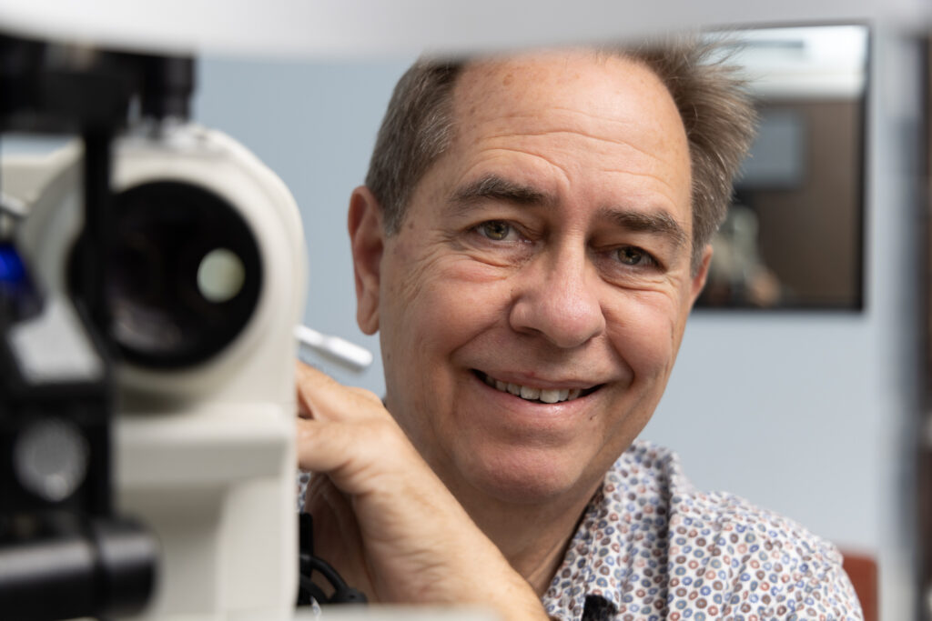 Dr Laurence Sullivan, a smiling man in a patterned shirt, sits behind an eye examination machine, looking directly at the camera in a clinical setting.