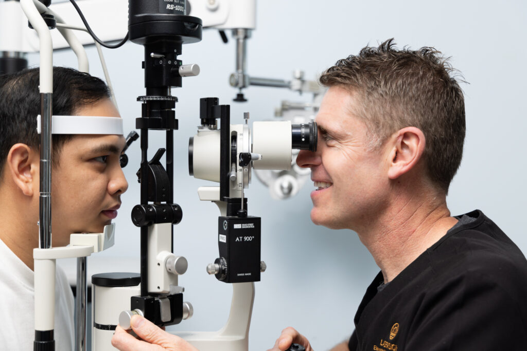 Dr Tim Steele examines a patient’s eyes using a slit lamp microscope in a clinical setting. The patient sits still whilst Dr Steele looks through the instrument, carefully focussing on the patient’s eyes.