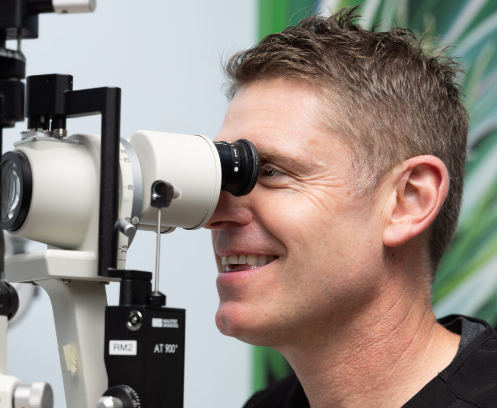 Dr Tim Steele in black scrub, smiles whilst looking through a slit lamp microscope during an eye examination, with a blurred green and blue background.
