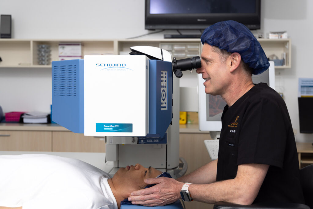 Dr Tim Steele, an eye doctor wearing a hairnet, examines a patient lying under a SCHWIND eye laser machine, preparing for an eye procedure in a clinical setting.