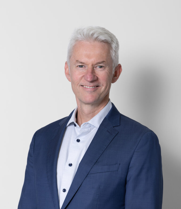 Dr Dermot Cassidy, a man with short white hair, stands smiling against a plain light background, dressed in a blue suit jacket over a light-coloured button-up shirt.
