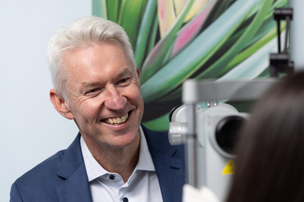 Dr Dermot Cassidy, a smiling man in a blue blazer, sits at an eye examination machine facing a person being tested; a large green abstract painting is visible in the background.