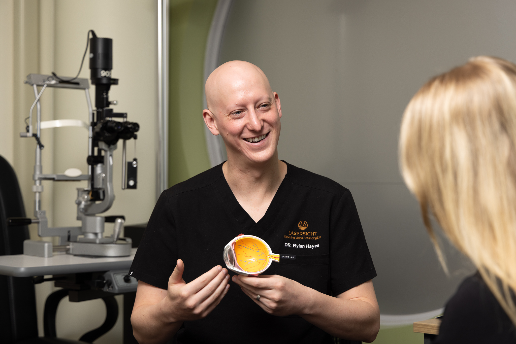 Dr Rylan Hayes, a smiling optometrist in black scrubs, holds a model of an eye whilst explaining something to a patient. An eye examination machine is visible in the background.