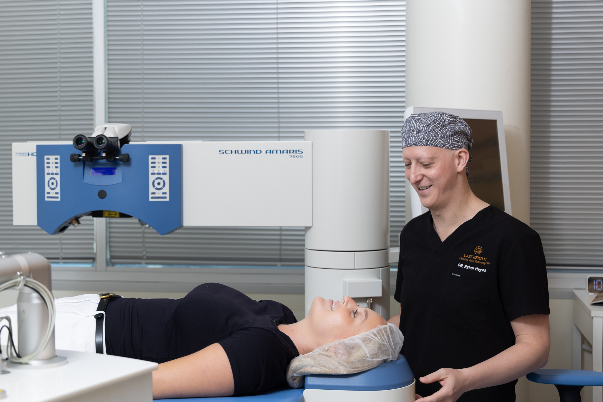A woman lies on a medical bed wearing a hair cover, preparing for an eye procedure. Dr Rylan Hayes, smiling in scrubs and a theatre cap, stands beside her next to a laser eye surgery machine in the modern clinic.