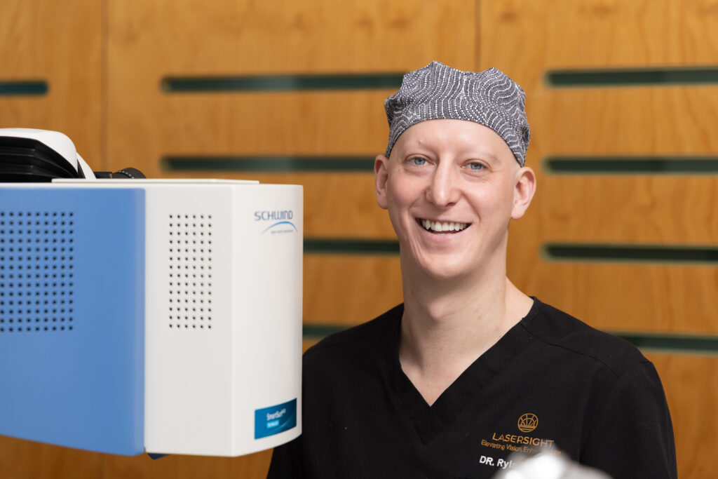 A smiling doctor, Dr Rylan Hayes, wearing a black scrub top and a patterned surgical cap stands beside a medical device against a wooden background. His shirt features a logo and the name Dr R.Y. embroidered on it.