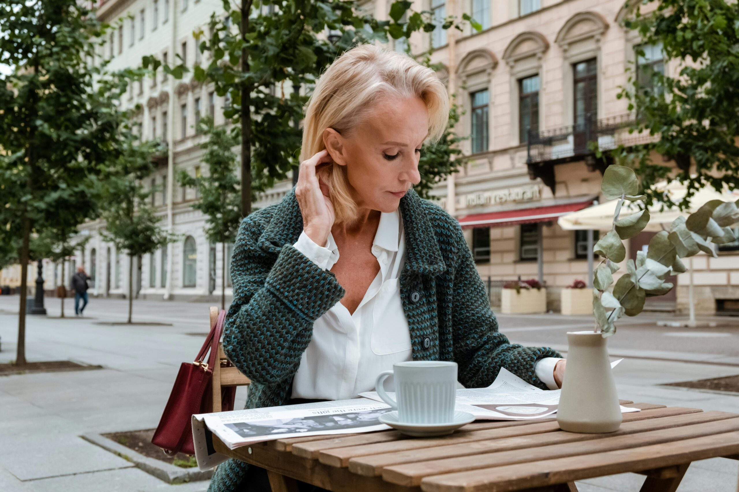 A woman with blonde hair sits at an outdoor café table, reading a newspaper with restored vision after presbyopia treatment. She wears a green cardigan, and a red bag hangs on her chair. Trees and buildings line the street behind her.