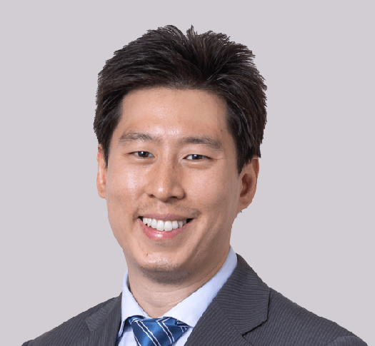 Dr Tony Chen, in a grey suit and blue striped tie, stands against a white background, smiling with his hands clasped in front of him.
