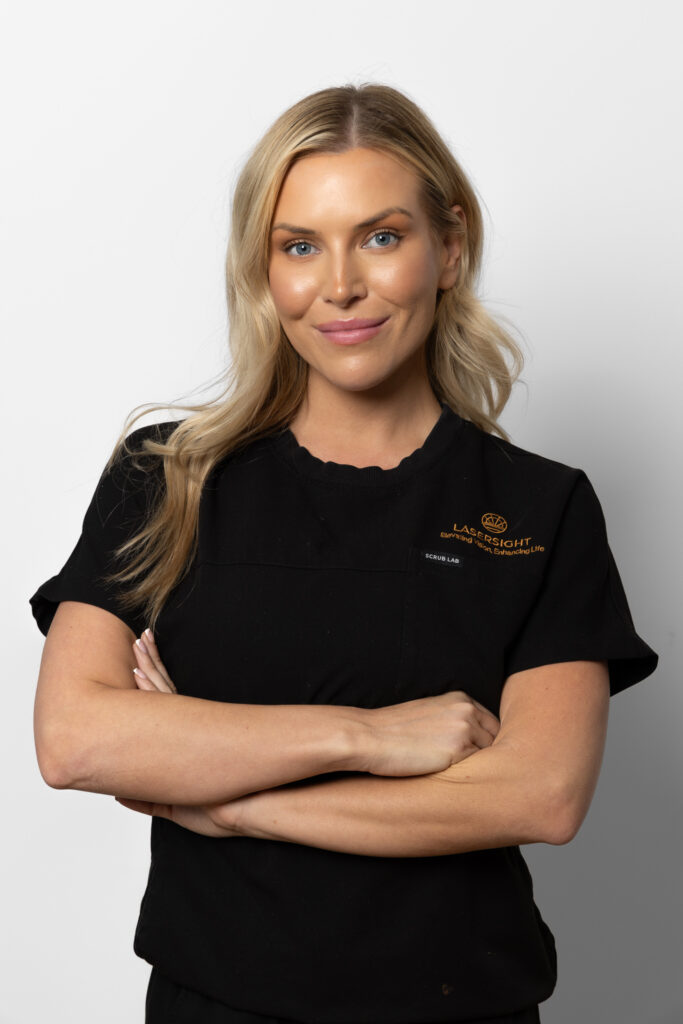 Ashleigh with long blonde hair wearing a black shirt with embroidered text stands against a plain white background, smiling with her arms folded.