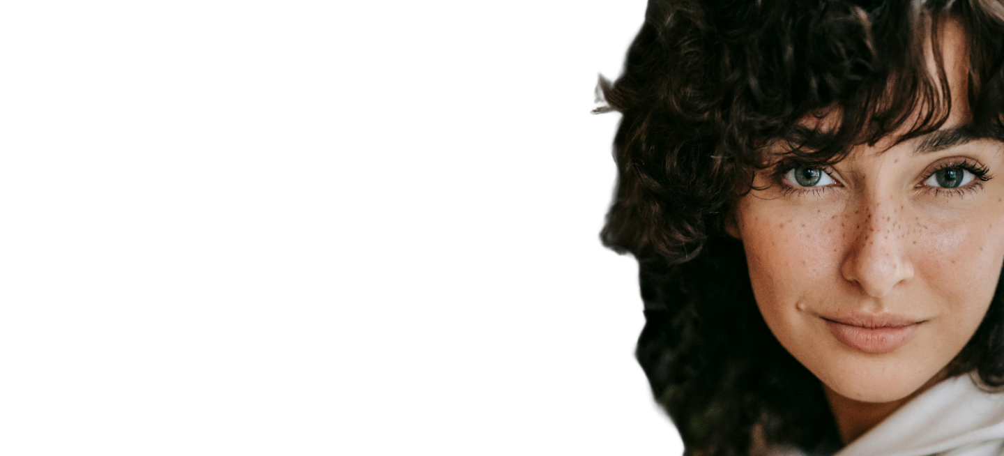 Close-up of a woman with curly dark hair, light skin, and freckles, smiling softly. The background is plain white, and only the right side of her face and hair are visible.