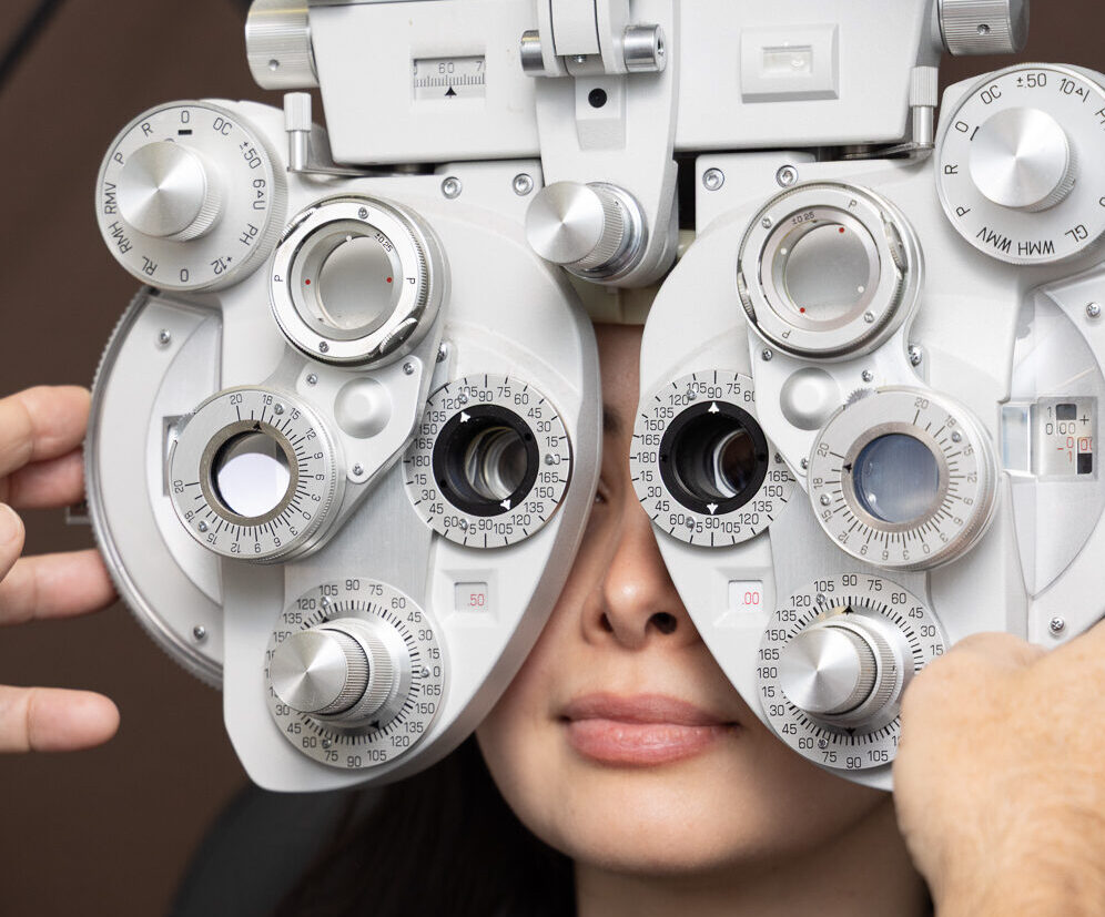 A woman is sitting for an eye test while an optometrist adjusts a phoropter in front of her face to test her vision.