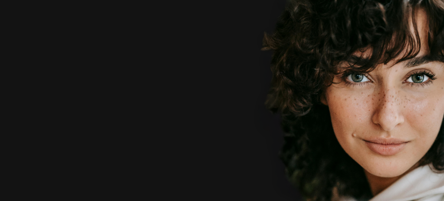 A woman with curly dark hair, green eyes, and freckles is smiling softly at the camera. Only the right side of her face is visible, with a dark background filling the left side of the image.