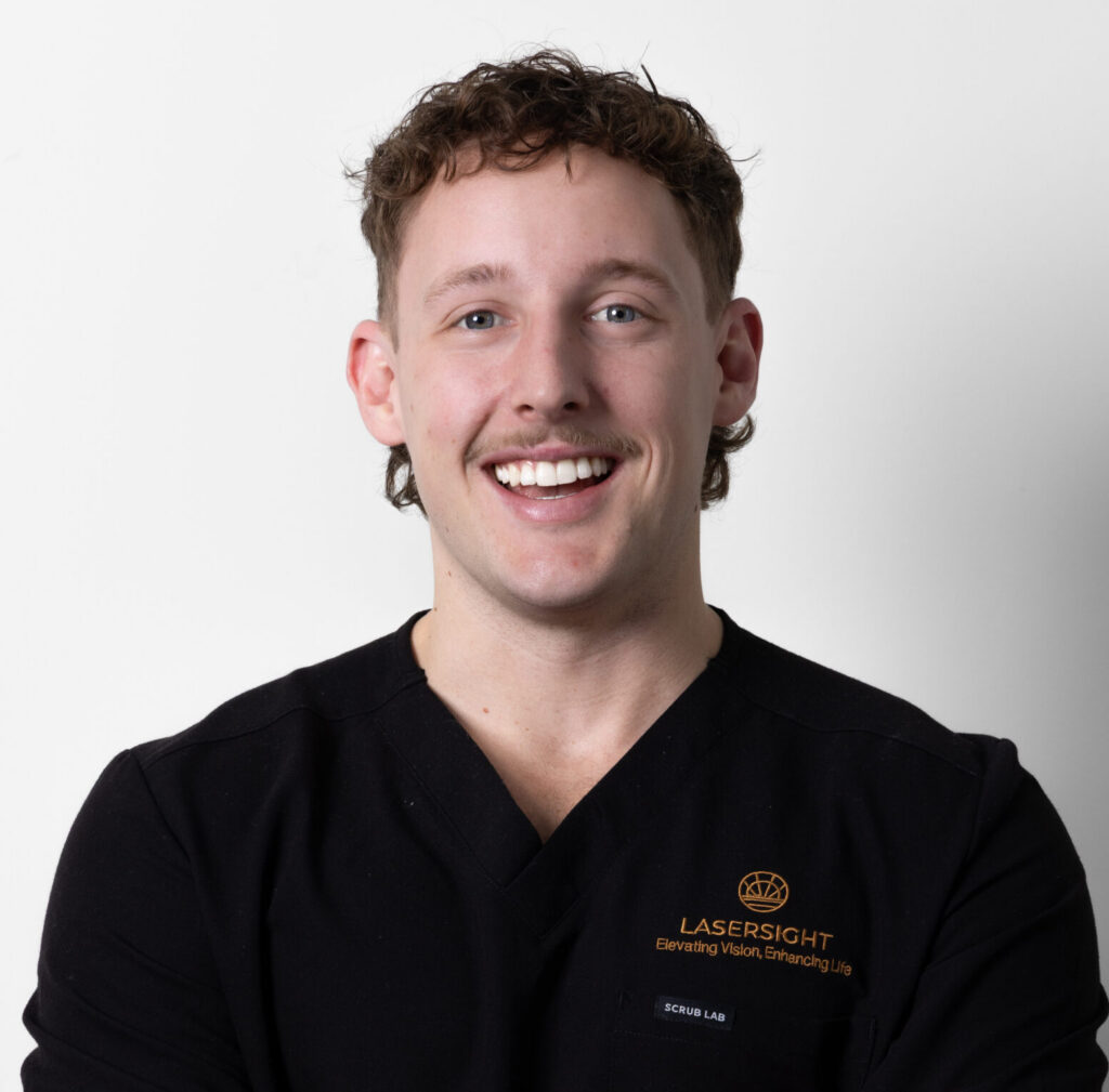 Bailey Hillier with short curly hair and a moustache smiles at the camera. He is wearing a black Lasersight scrub top, standing against a plain white background, ready to assist patients at Mount Coolum.