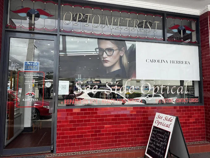 64248d3a-a5dc-44ab-9e67-dc50e459d6f0 Shopfront of See Side Optical in LASERSIGHT Thirroul with a large window advert featuring a woman in glasses, red tiled exterior, and an OPEN sign on the door. A pavement sign with shop info stands outside.