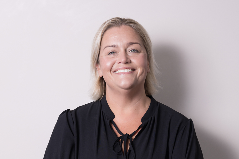 A woman with short blonde hair smiles at the camera, wearing a black blouse with a tie at the neckline, standing in front of a plain light-coloured background at LASERSIGHT Thirroul.
